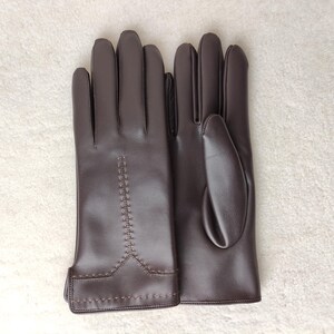 Puede incluir: Un par de guantes de cuero marr&oacute;n. Los guantes son de color marr&oacute;n oscuro liso con un dise&ntilde;o cosido en el dorso de la mano. Los guantes est&aacute;n planos sobre una superficie blanca.