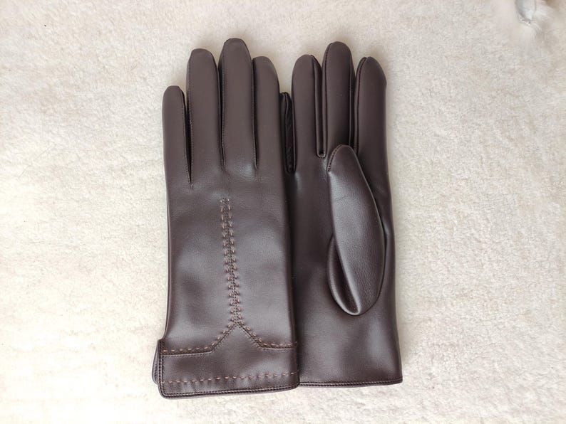 Puede incluir: Un par de guantes de cuero marr&oacute;n. Los guantes son de color marr&oacute;n oscuro liso con un dise&ntilde;o cosido en el dorso de la mano. Los guantes est&aacute;n planos sobre una superficie blanca.