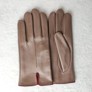 May include: A pair of smooth, light brown leather gloves featuring a vertical seam on the back of the hand and a small slit at the wrist. The interior lining of the gloves is visible at the cuff, showing a vibrant red color.
