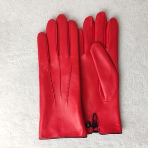 May include: A pair of bright red leather gloves featuring three decorative stitched lines on the back of the hand and a small black button closure at the wrist. The gloves have a smooth finish and a clean, structured design for the hands.