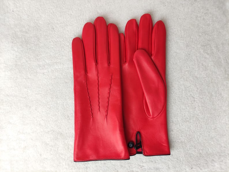 May include: A pair of bright red leather gloves featuring three decorative stitched lines on the back of the hand and a small black button closure at the wrist. The gloves have a smooth finish and a clean, structured design for the hands.