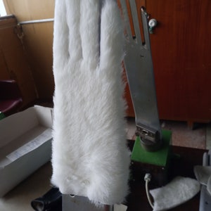 May include: A white, furry glove, possibly a costume piece, is mounted on a metal hand form. The glove has a soft, plush texture and covers the entire hand. The metal hand has a silver finish and is attached to a green base.