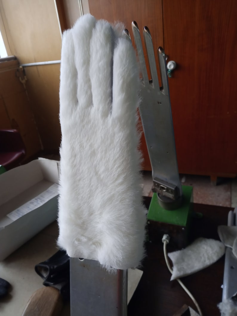May include: A white, furry glove, possibly a costume piece, is mounted on a metal hand form. The glove has a soft, plush texture and covers the entire hand. The metal hand has a silver finish and is attached to a green base.