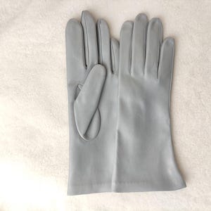 May include: A pair of light gray leather gloves with a smooth finish. The gloves feature a classic design with four fingers and a thumb section, extending to the wrist. The material appears soft and uniform in color throughout the surface.