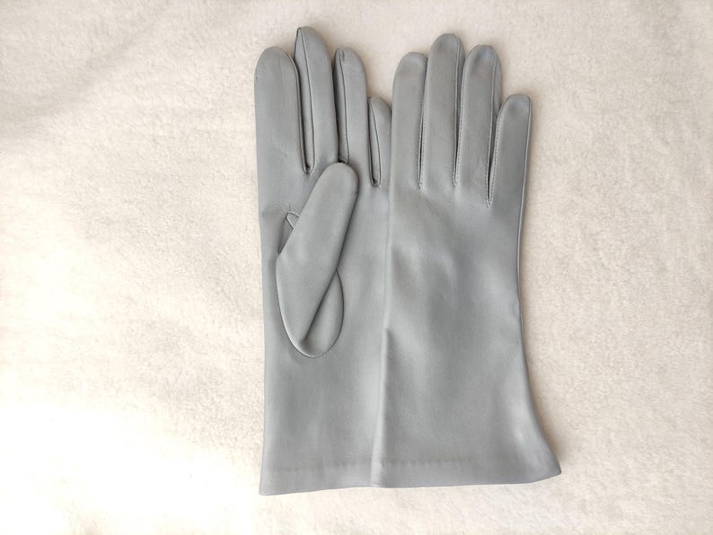 May include: A pair of light gray leather gloves with a smooth finish. The gloves feature a classic design with four fingers and a thumb section, extending to the wrist. The material appears soft and uniform in color throughout the surface.