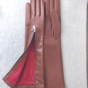 May include: A pair of brown leather gloves featuring a silver-toned metal zip along the side of the wrist. The interior of the left glove is lined with a vibrant red fabric, which is visible when the zip is partially opened.