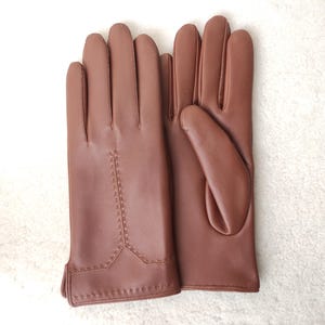 Puede incluir: Un par de guantes de cuero marr&oacute;n. Los guantes son de color liso con un detalle de costura en la parte posterior de la mano. Los guantes se muestran sobre una superficie blanca.