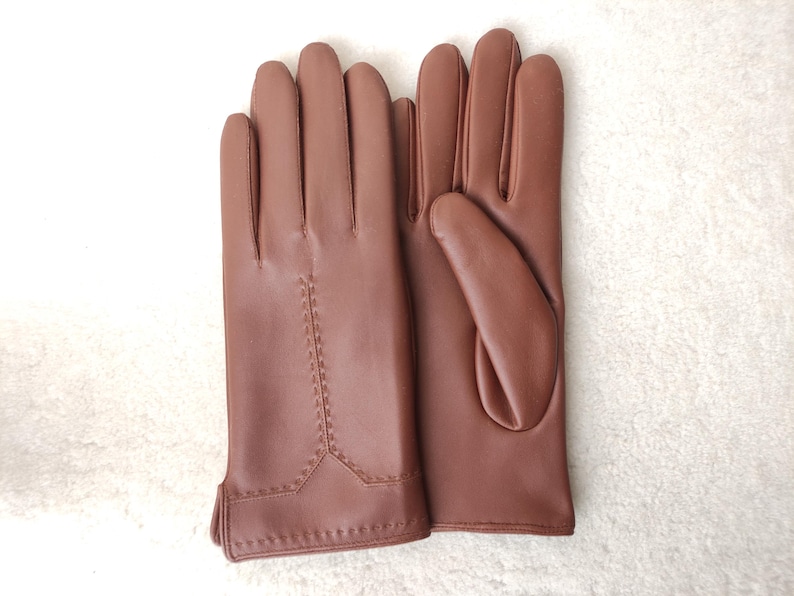 Puede incluir: Un par de guantes de cuero marr&oacute;n. Los guantes son de color liso con un detalle de costura en la parte posterior de la mano. Los guantes se muestran sobre una superficie blanca.