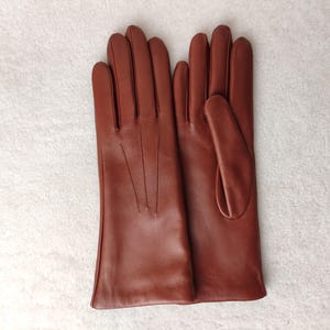 Peut inclure: Une paire de gants en cuir marron moyen lisse, dot&eacute;e de trois lignes de coutures d&eacute;coratives verticales sur le dos de la main. Les gants pr&eacute;sentent une coupe classique avec un pouce et quatre doigts, offrant une finition uniforme.