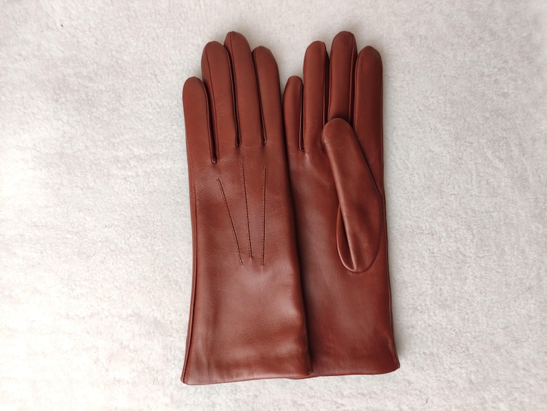 Peut inclure: Une paire de gants en cuir marron moyen lisse, dot&eacute;e de trois lignes de coutures d&eacute;coratives verticales sur le dos de la main. Les gants pr&eacute;sentent une coupe classique avec un pouce et quatre doigts, offrant une finition uniforme.