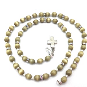 Può includere: Una collana di perline verdi e bianche con un ciondolo a forma di croce d'argento. Le perline sono di forma ovale e hanno un leggero luccichio.