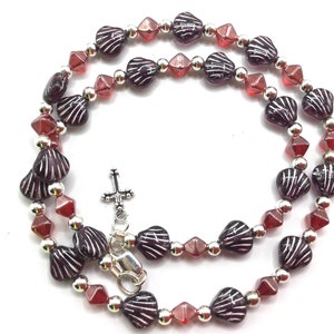 May include: A dark red and silver beaded necklace with a small silver cross charm. The beads are shaped like seashells and have a textured surface.