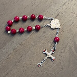 May include: A red beaded rosary with a silver crucifix and a silver charm featuring a depiction of the Virgin Mary.