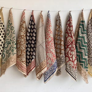 Può includere: Una collezione di dieci bandane decorate appese a una corda. Le bandane presentano vari disegni, tra cui paisley, floreali e geometrici, in colori come marrone, nero, rosso e verde. Ogni bandana è piegata e presentata su uno sfondo bianco.