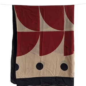 May include: A rectangular textile with a geometric pattern of red semi-circles and beige squares. Large black circles are at the bottom, with a black border. The textile is hanging from a thin wire against a white background.