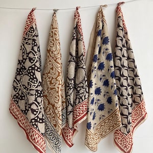 May include: Five patterned scarves hanging on a wire. The scarves feature various designs in colors like brown, red, and blue. Each scarf has a patterned border. The scarves are made of a lightweight fabric.