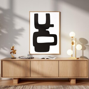 Puede incluir: Un cuadro abstracto en blanco y negro enmarcado cuelga sobre un mueble de madera clara. Una lámpara dorada con bombillas esféricas y una pequeña escultura dorada añaden toques decorativos. La habitación tiene una estética moderna y minimalista.