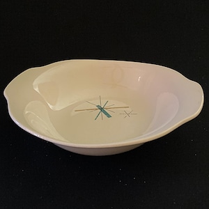 May include: A cream-colored, mid-century modern serving bowl with a unique, asymmetrical shape. The bowl features a stylized turquoise and gold starburst design in the center. The bowl is made of ceramic.