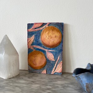 May include: A ceramic tile with a blue background and a raised design of pink leaves and orange fruit. The tile has a textured surface and a rustic look.