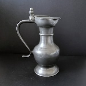 May include: A pewter pitcher with a hinged lid and a curved handle. The pitcher has a bulbous body and a round base.