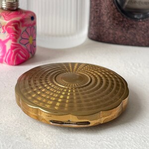Peut inclure: Un compact de couleur or avec un motif circulaire en relief. Le compact a un bord festonné et un petit loquet rectangulaire.