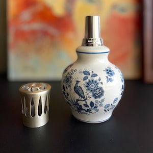 May include: A white ceramic oil lamp with blue floral and bird designs. It has a silver-colored metal top and a separate metal diffuser. The lamp is on a dark surface, with a colorful painting in the background.