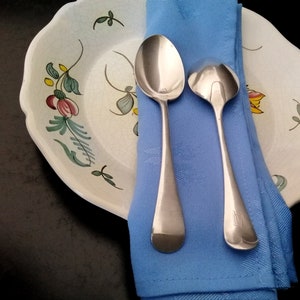 May include: Two silver spoons on a blue napkin, with a white and green floral patterned plate in the background.