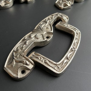 Puede incluir: Tiradores de cajón de metal plateado con un diseño floral ornamentado. Los tiradores tienen una forma rectangular con una parte superior e inferior curvada.