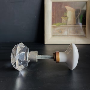 Peut inclure: Une poignée de porte en cristal avec un design facetté et une poignée en porcelaine blanche à l'autre extrémité. La poignée est fixée à une tige métallique.