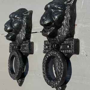 Op de afbeelding: Een paar zwarte gietijzeren deurkloppers in de vorm van een leeuwenkop met sierlijke ringen. De leeuwenkoppen zijn gedetailleerd met getextureerd haar en hebben open monden. De ringen zijn rond en hebben een decoratief ontwerp.