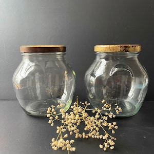 Könnte beinhalten: Zwei durchsichtige Glasbehälter mit goldenen Deckeln. Die Behälter sind leer und stehen auf einer schwarzen Oberfläche. Ein Zweig getrockneter weißer Blumen liegt vor den Behältern.