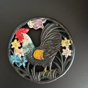 Puede incluir: Un posavasos de hierro fundido con un gallo colorido rodeado de flores. El gallo es negro, blanco, rojo, amarillo y verde. Las flores son rosas, azules, amarillas y verdes.