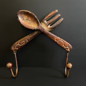May include: A rustic brown metal spoon and fork wall hook with decorative swirls. The hooks are in the shape of a spoon and fork, crossed over each other.