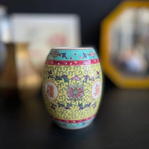 Puede incluir: Un pequeño jarrón de cerámica amarillo con un diseño floral y caracteres chinos. El jarrón tiene una forma redondeada y un borde turquesa.