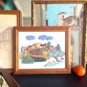 Peut inclure: Une œuvre d'art encadrée représentant un paysage avec une maison blanche, des arbres et des nuages bleus. L'œuvre est réalisée avec divers morceaux de tissu verts, bruns et roses. Le cadre est en bois.