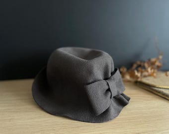 Pode incluir: Um chapéu de feltro cinza escuro com um grande laço de lado. O chapéu tem uma aba larga e uma coroa arredondada.