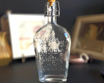 May include: A clear glass bottle with a cork stopper and a metal wire closure. The bottle has a floral design and the word "Frau" etched into the glass.