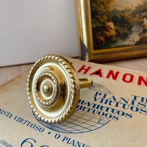 Puede incluir: Un pomo de metal dorado con un diseño de cuerda alrededor del borde. El pomo tiene un centro elevado con un círculo más pequeño en el medio.