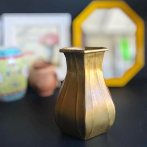 Puede incluir: Un jarrón de metal dorado con base cuadrada y diseño acanalado. El jarrón está sobre una superficie negra.