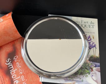 Peut inclure: Un miroir rond, encadré d'argent, avec une surface réfléchissante. Le miroir est posé sur un livre avec le texte "vrez l'art du UQUET Simon Lycett" sur la couverture.