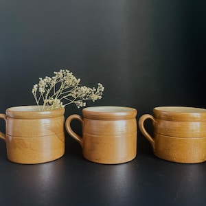May include: Three brown ceramic mugs with a rustic, handmade look. The mugs have a rounded shape and a slightly flared lip.