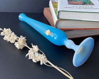 May include: A light blue glass vase with a wide base and a label that says "Bohemian". The vase is sitting on a stack of books. A dried flower stem is lying on a black surface.