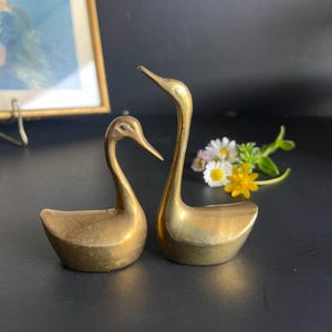 May include: Two brass swan ornaments. One swan has its neck extended upwards, the other has its neck curved downwards. The swans are positioned next to each other. The ornaments are on a dark surface, with a framed picture and flowers in the background.