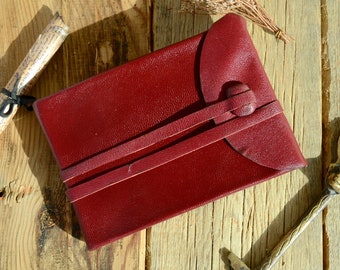 May include: A burgundy leather journal with a flap closure and a leather strap with a button. The journal is lying on a wooden surface.