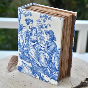 May include: A handcrafted journal with a blue and white toile cover depicting a rural scene. The pages are aged, and the book is open, showing the textured paper. A feather lies close by.