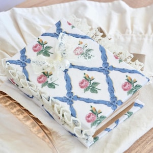 May include: A small, blue and white floral patterned journal with a lace trim and a white pearl embellishment. The journal is closed and lying on a white fabric surface.