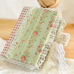 Puede incluir: Un pequeño diario de tela verde y rosa con ribete de encaje. El diario tiene un patrón floral y está atado con una cinta de encaje blanca.