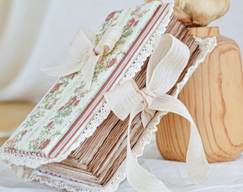 May include: A vintage-style journal with a floral pattern cover in soft green and red. The journal is tied with a cream-colored ribbon and features a lace trim. The pages are aged, giving it a classic, antique look.