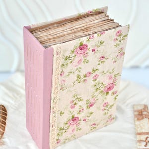 Pode incluir: Um diário feito à mão com lombada rosa e capa floral com rosas cor-de-rosa e folhas verdes sobre um fundo creme. As páginas do diário são visíveis, e uma borda de renda decorativa contorna o padrão floral.