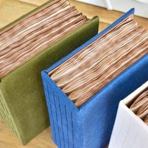 Può includere: Tre libri fatti a mano con copertine strutturate in verde, blu e bianco. Le pagine sono invecchiate con una calda tonalità marrone. I libri sono disposti su una superficie di legno chiaro, mostrando la loro unica qualità artigianale.